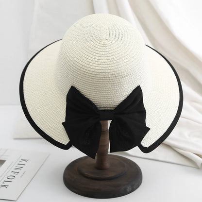 White straw hat with black edge trim and oversized bow, displayed elegantly on wooden stand.